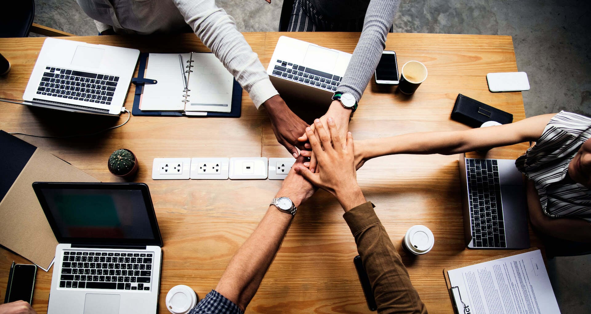 Overhead shot of 5 people putting hands in middle of conference table to show teamwork